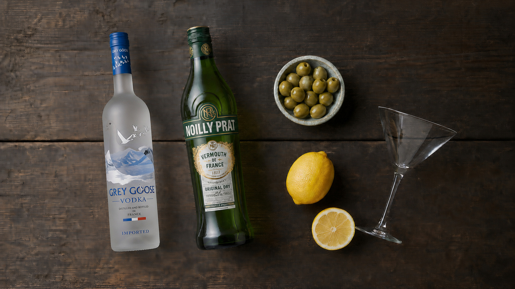 Flatlay mit Grey Goose Wodka, Noilly Prat Wermut, Oliven, Zitrone und Martiniglas auf dunklem Holz